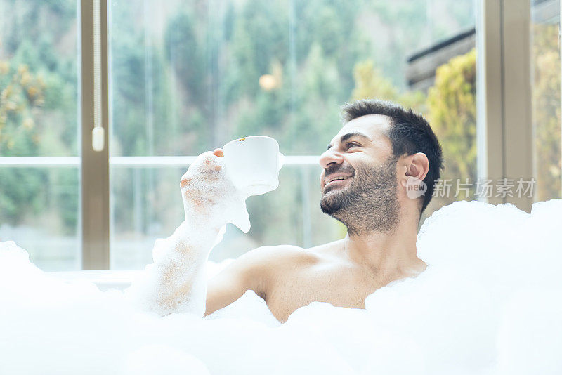 一个男人微笑着，拿着一杯咖啡/茶在一个热水浴缸里望着外面的森林景色